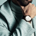 Un homme vêtu d'un manteau gris clair et d'un col roulé pose avec sa main sur son menton, montrant une montre-bracelet avec un cadran blanc et un bracelet noir.