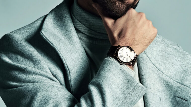 Un homme vêtu d'un manteau gris clair et d'un col roulé pose avec sa main sur son menton, montrant une montre-bracelet avec un cadran blanc et un bracelet noir.