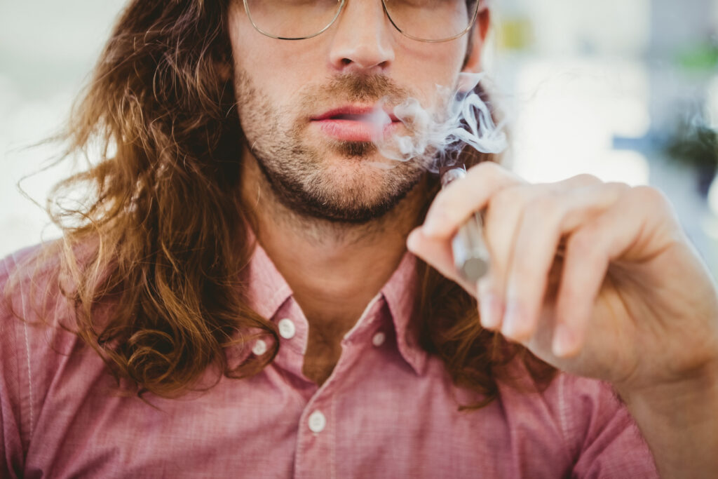 Une personne aux cheveux longs et aux lunettes, vêtue d'une chemise rose, tient un dispositif de vaporisation et expire de la vapeur.