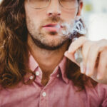 Une personne aux cheveux longs et aux lunettes, vêtue d'une chemise rose, tient un dispositif de vaporisation et expire de la vapeur.