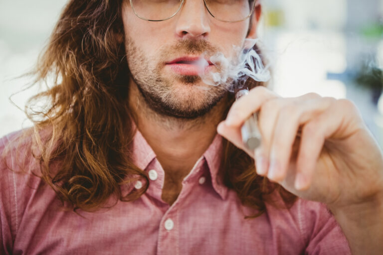 Une personne aux cheveux longs et aux lunettes, vêtue d'une chemise rose, tient un dispositif de vaporisation et expire de la vapeur.